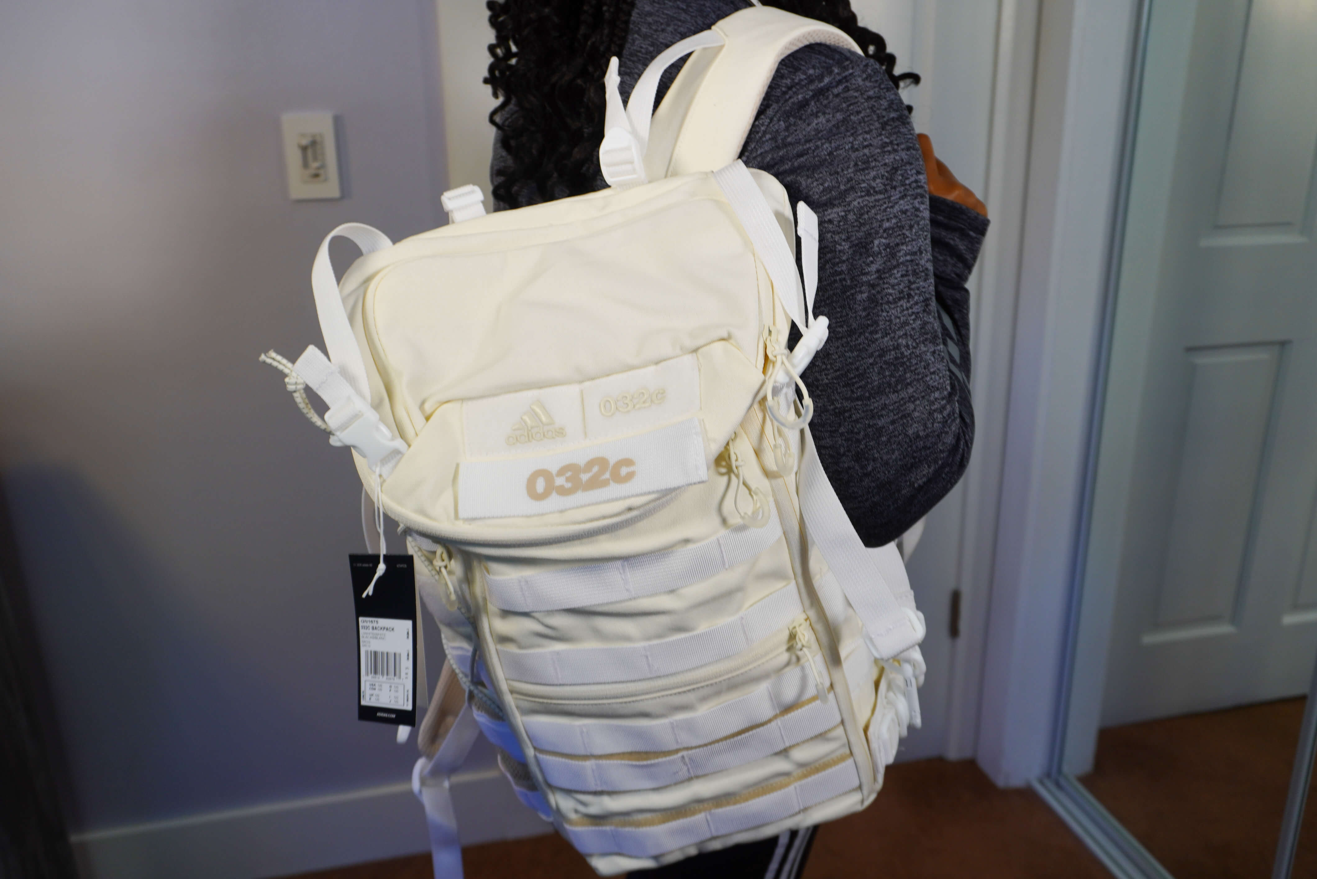 Image of me holding the backpack on one shoulder. The bag is a cream color with white straps. On the front it says Adidas and 032c.