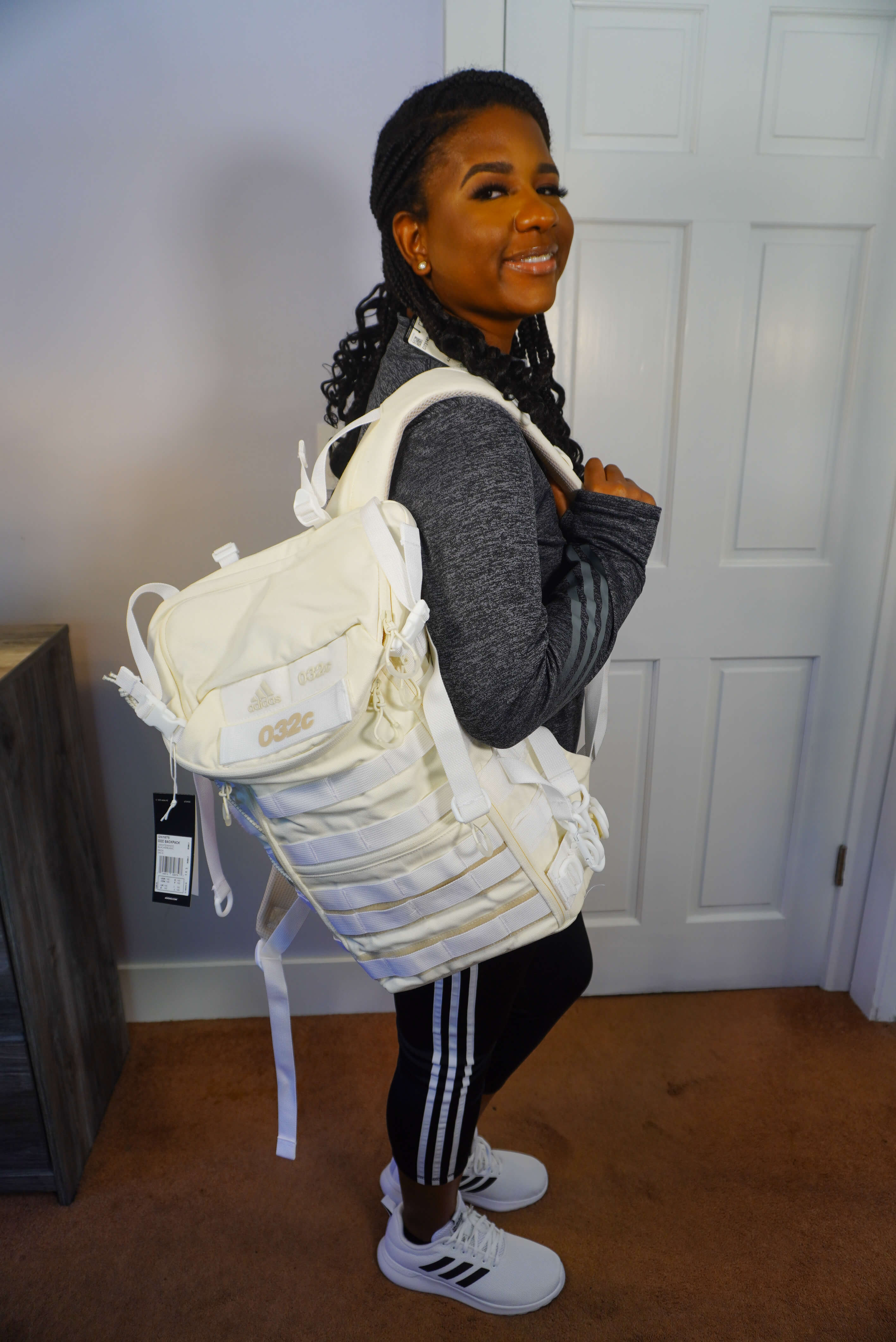 Image of me posing with a cream Adidas backpack. I am wearing white Adidas sneakers, with black leggings, and a long sleeve gray top.