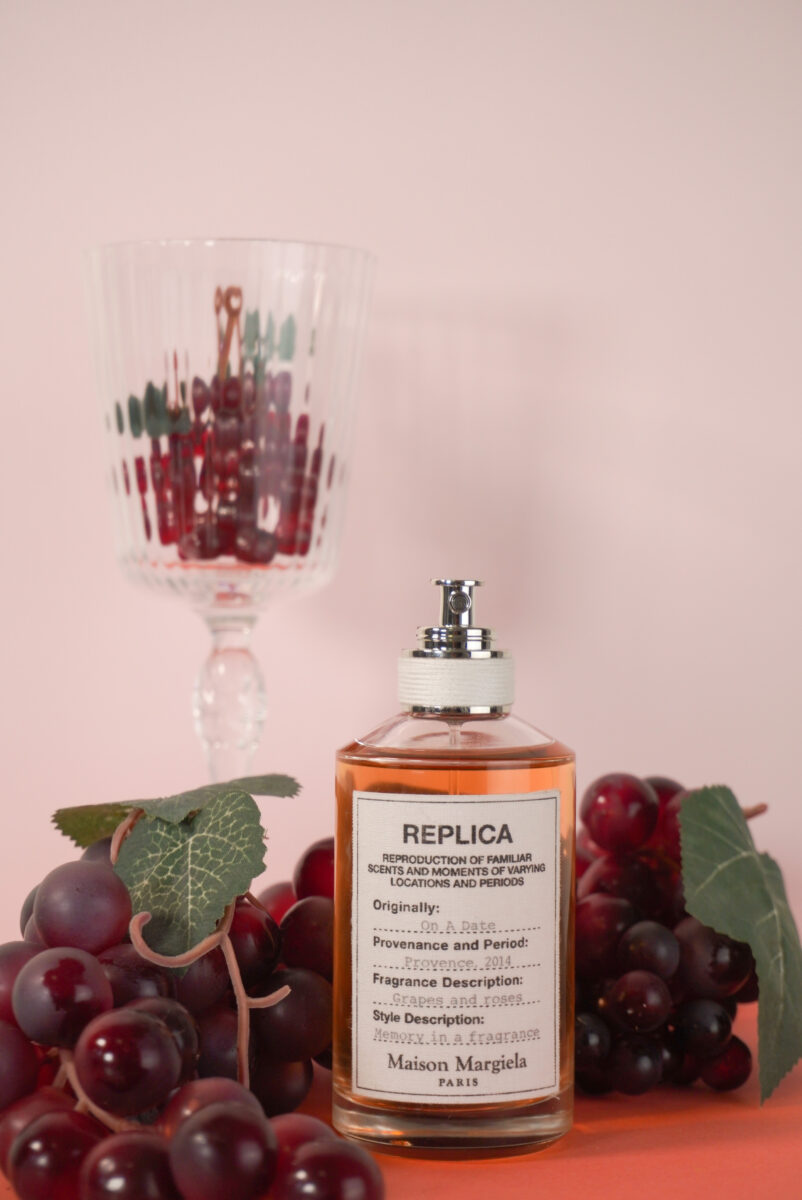 Image of the MM Replica fragrance surrounded by grapes with a wine glass in the back.