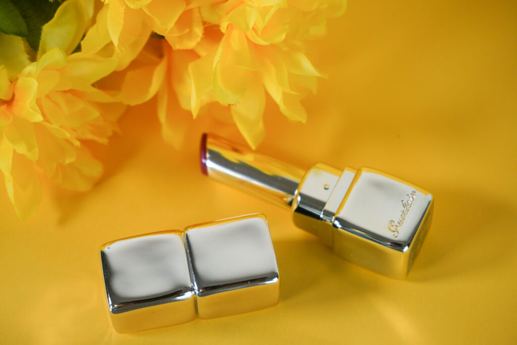 Image of Guerlain Kiss Kiss Shine Bloom lipstick with a flower in the background