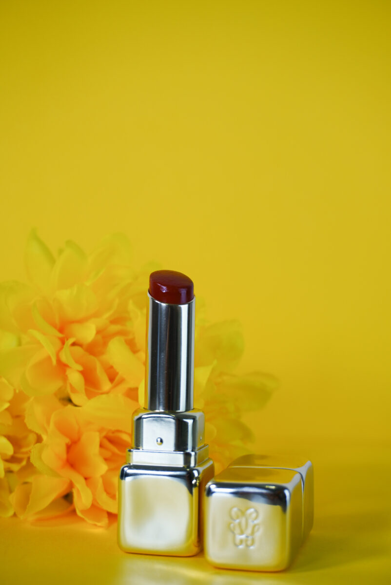 Image of Guerlain's Kiss Kiss Shine Bloom lipstick with the cap off and a yellow flower in the background